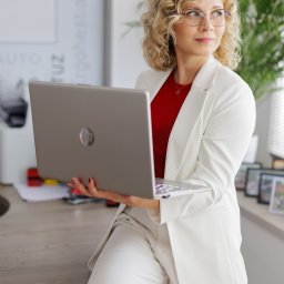 SALVO LEASINGI I UBEZPIECZENIA - Kobieta z kręconymi blond włosami i w okularach, w białym garniturze i czerwonej bluzce, siedzi na biurku z otwartym srebrnym laptopem HP, patrząc w prawo. W tle zielona roślina i ramki na parapecie.