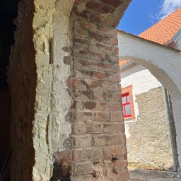 BARTOSZ SUDAK - Siła Piasku - Fragment muru z cegły z widoczną pianką montażową i śladami czyszczenia, w tle budynek z czerwoną stolarką okienną i fragmentem nieba. Prace przygotowawcze do czyszczenia.