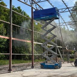 BARTOSZ SUDAK - Siła Piasku - Prace na konstrukcji stalowej z użyciem podnośnika Genie GS 3246. Widoczne efekty czyszczenia, mgła wodna. W tle zieleń drzew i błękitne niebo.
