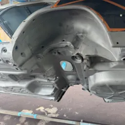 Podwozie samochodu po piaskowaniu, zabezpieczone pomarańczową taśmą. Elementy zawieszenia i nadkola w szarym podkładzie. Widoczna konstrukcja nośna podtrzymująca karoserię.