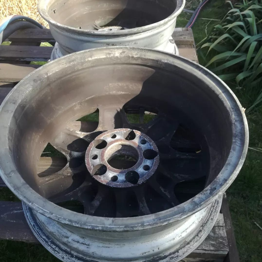 Dwie aluminiowe felgi samochodowe przed czyszczeniem, widoczne ślady korozji i zabrudzeń na powierzchni, ustawione na drewnianej palecie na zewnątrz.