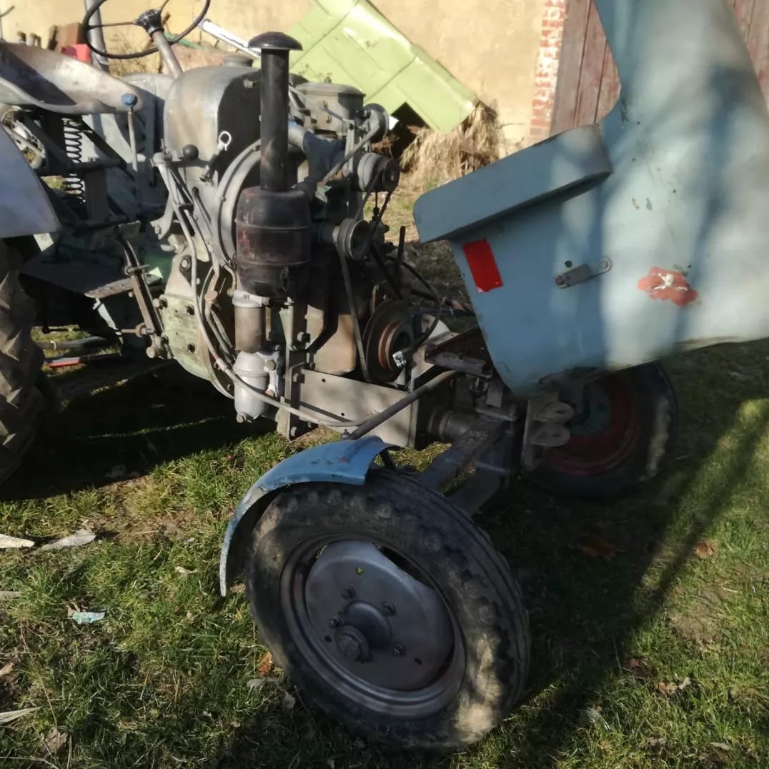 Stary, zaniedbany traktor rolniczy z widocznymi elementami silnika, stojący na trawie w słoneczny dzień. Widoczne ślady rdzy i zużycia.