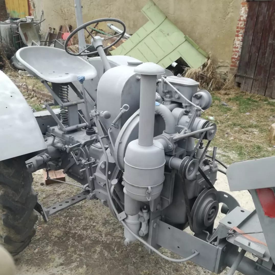 Szary, odrestaurowany traktor rolniczy z widocznym silnikiem, na tle muru budynku i zielonej konstrukcji, prezentujący efekt po renowacji.