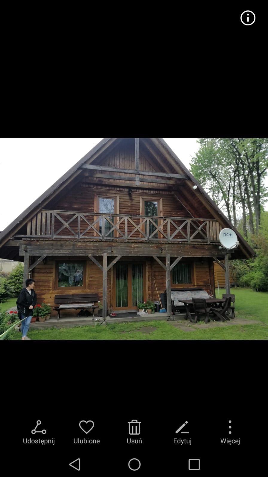 Drewniany dom z balkonem i satelitą na tle zieleni. Kobieta zamiata przed wejściem. Widoczne meble ogrodowe i kwiaty w donicach. Jasne, naturalne oświetlenie.