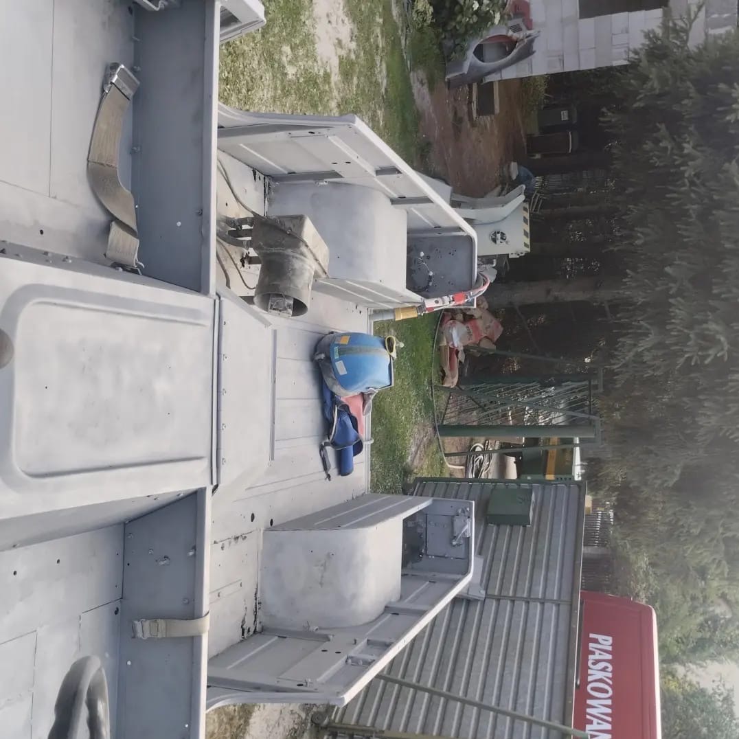 Karoseria pojazdu po piaskowaniu, przygotowana do dalszych prac lakierniczych. Widoczny hełm ochronny i elementy konstrukcyjne.