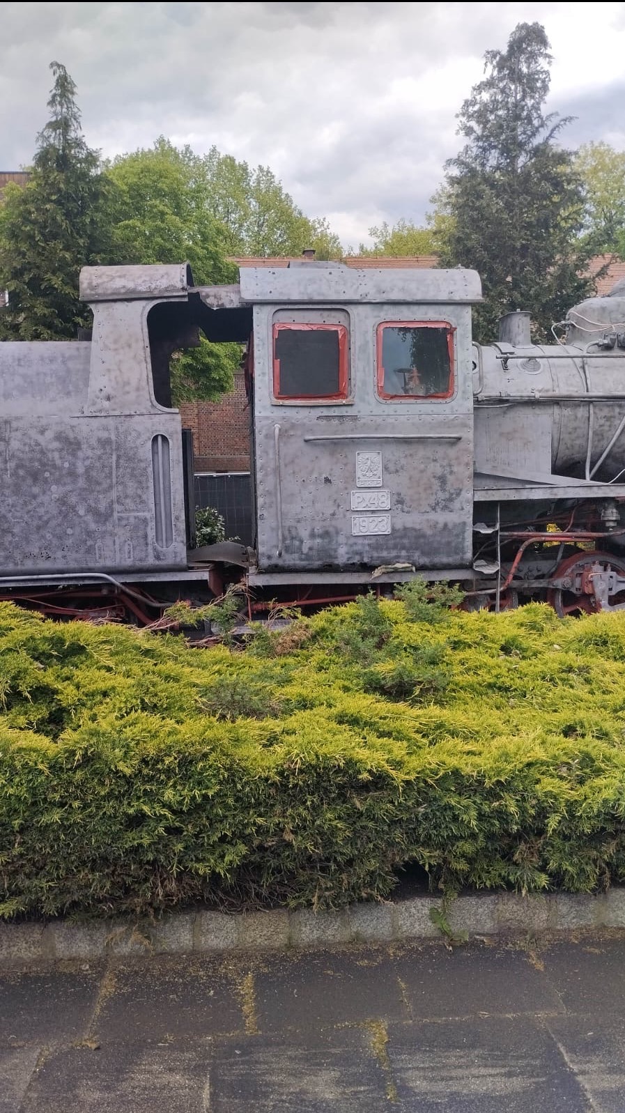 Lokomotywa Px48 z 1923 roku, eksponat muzealny, z widocznymi śladami korozji i upływu czasu, otoczona zielenią. Ujęcie z poziomu gruntu.
