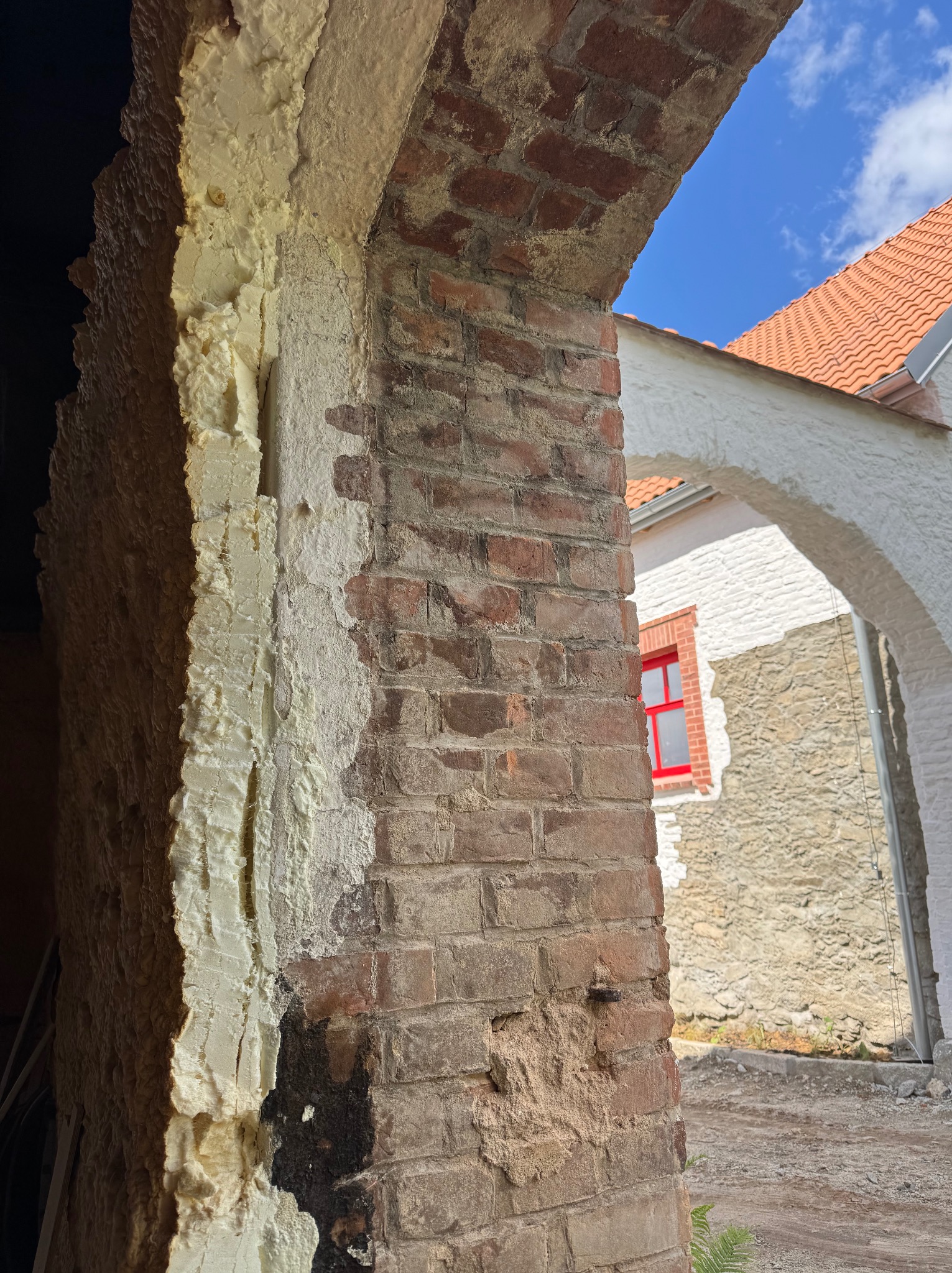 Fragment muru z cegły z widoczną pianką montażową i śladami czyszczenia, w tle budynek z czerwoną stolarką okienną i fragmentem nieba. Prace przygotowawcze do czyszczenia.