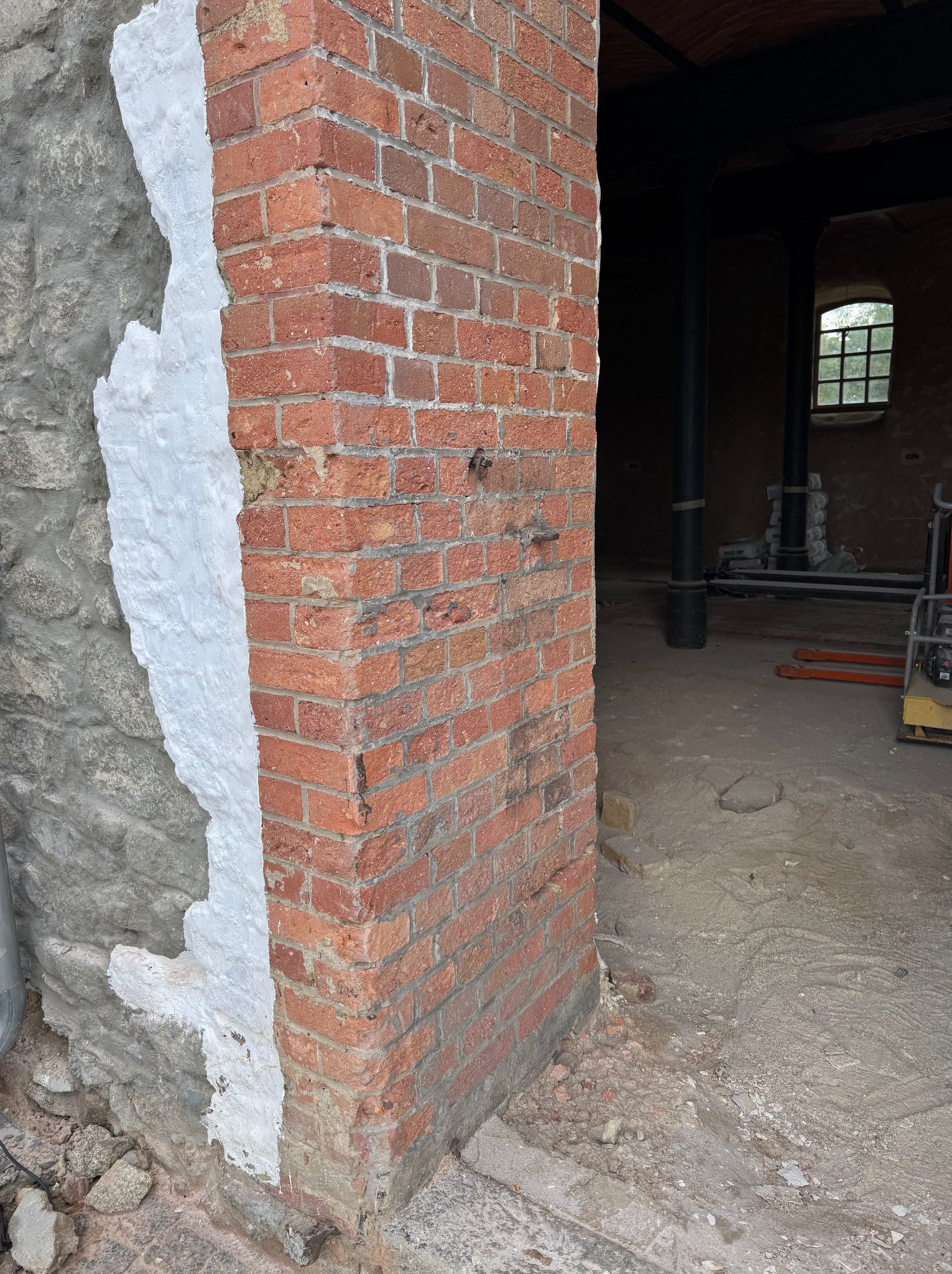 Fragment ściany z czerwonej cegły, częściowo oczyszczonej z białej farby, sąsiadującej z surowym kamieniem. Widoczne ślady prac renowacyjnych.