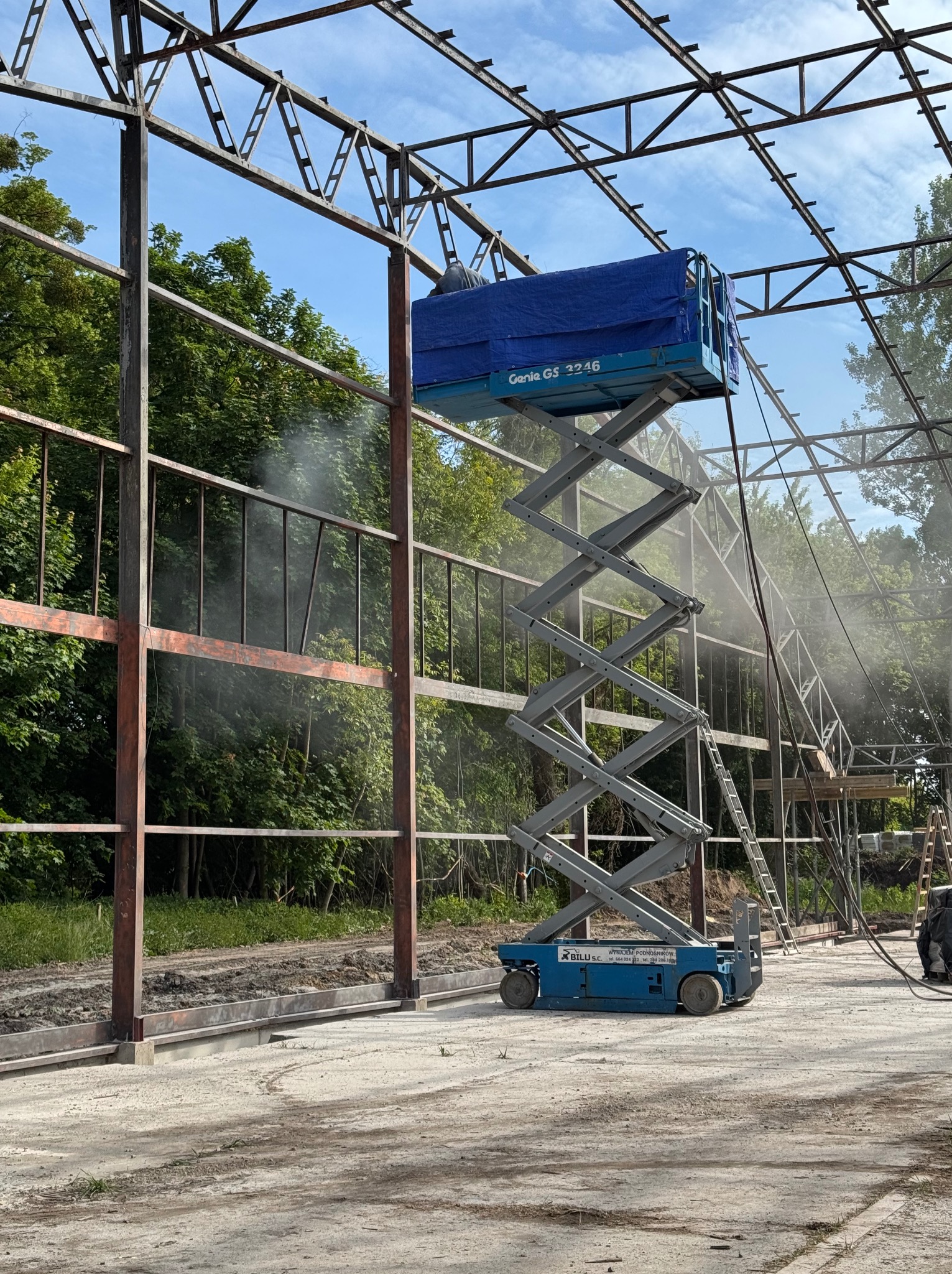 Prace na konstrukcji stalowej z użyciem podnośnika Genie GS 3246. Widoczne efekty czyszczenia, mgła wodna. W tle zieleń drzew i błękitne niebo.