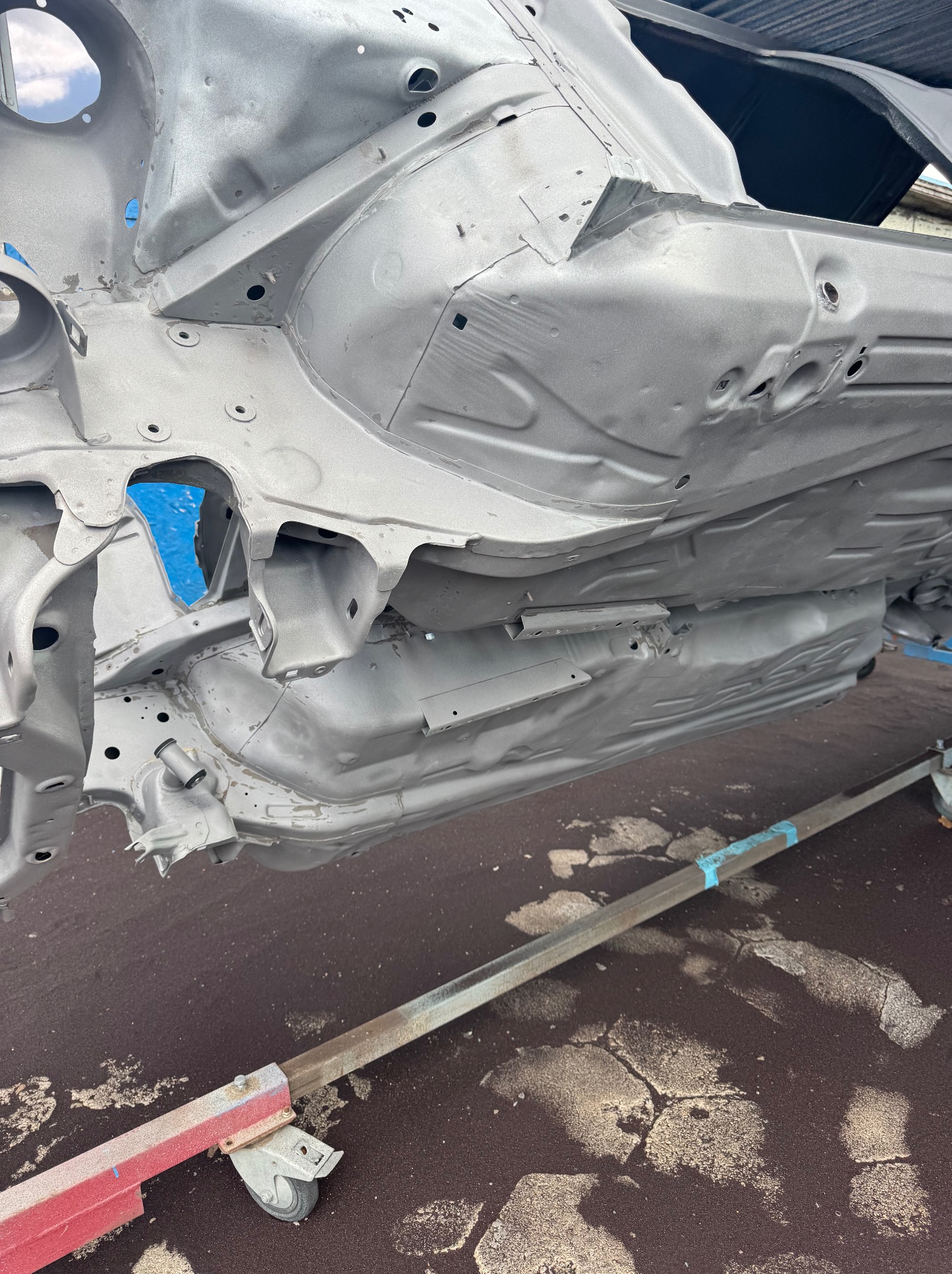 Szare podwozie samochodu po renowacji, widoczne od spodu, wsparte na stojaku. Niebieska taśma zabezpieczająca detale. Widoczne ślady szlifowania.