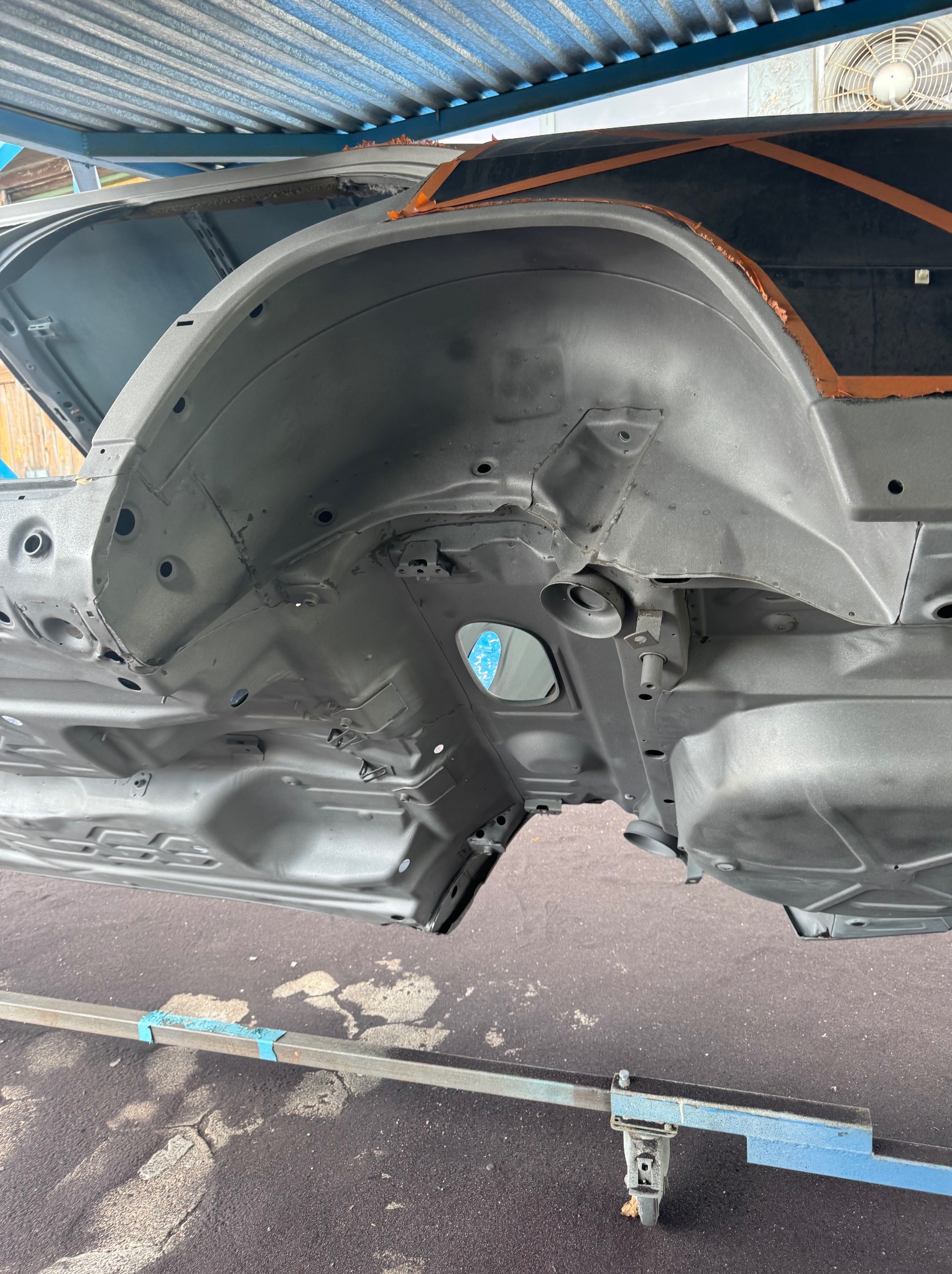 Podwozie samochodu po piaskowaniu, zabezpieczone pomarańczową taśmą. Elementy zawieszenia i nadkola w szarym podkładzie. Widoczna konstrukcja nośna podtrzymująca karoserię.