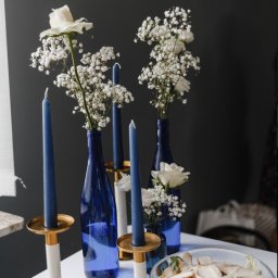 Zora Sp. Z o.o. - Dekoracja stołu z białymi kwiatami w niebieskich butelkach i granatowymi świecami w złotych świecznikach, obok miska z sałatką z kurczakiem na białym stole.