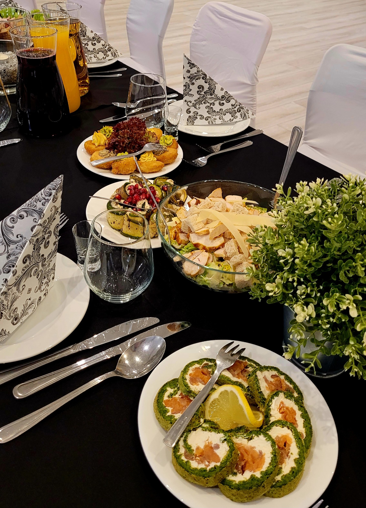 Elegancko nakryty stół z różnorodnymi daniami cateringowymi: sałatka Cezar, roladki szpinakowe z łososiem i przekąski. Czarny obrus, białe krzesła, dekoracyjne serwetki.