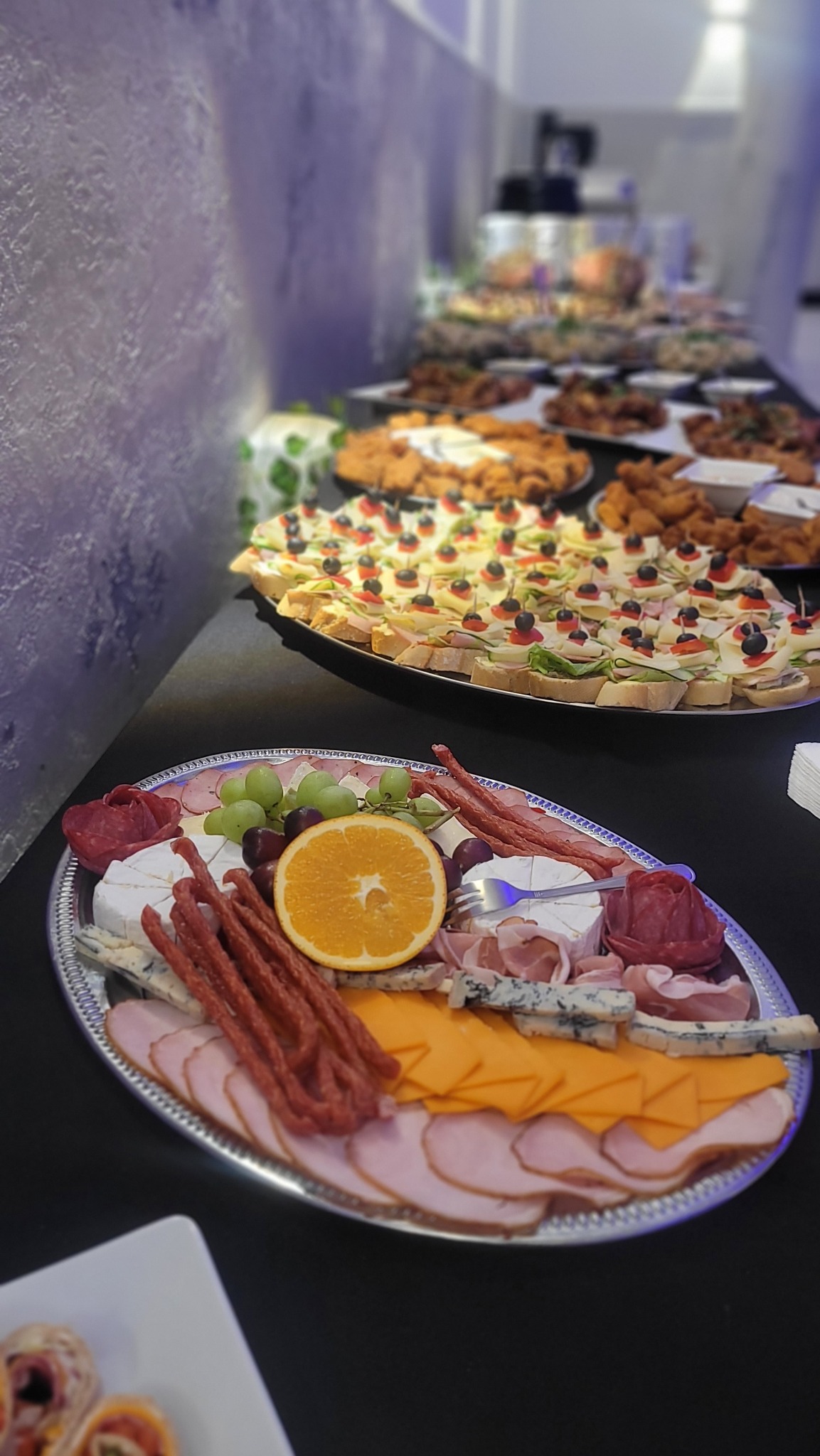 Srebrna patera z wędlinami, serami, owocami i pomarańczą na tle stołu z przekąskami. Kanapki na drugim planie. Catering na przyjęciu.