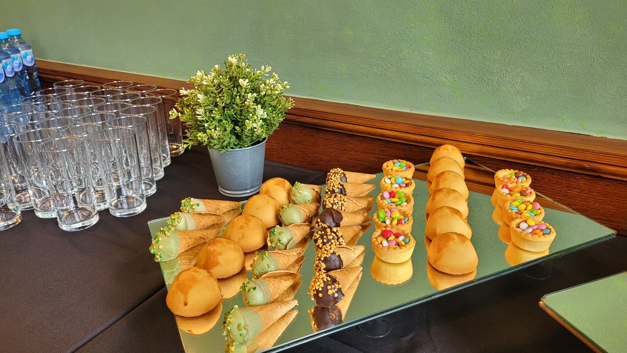 Słodki stół z deserami: mini tartaletki z kolorowymi cukierkami, rożki waflowe z kremem i kulki, ustawione na lustrzanej tacy obok szklanek i dekoracji roślinnej.