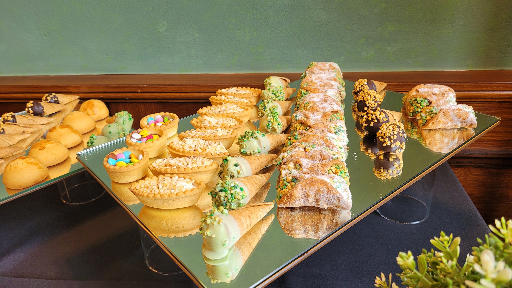 Elegancki bufet deserowy z różnorodnymi słodkościami: tartaletki z kremem, wafelki, cannoli posypane pistacjami i czekoladowe trufle na lustrzanej tacy.