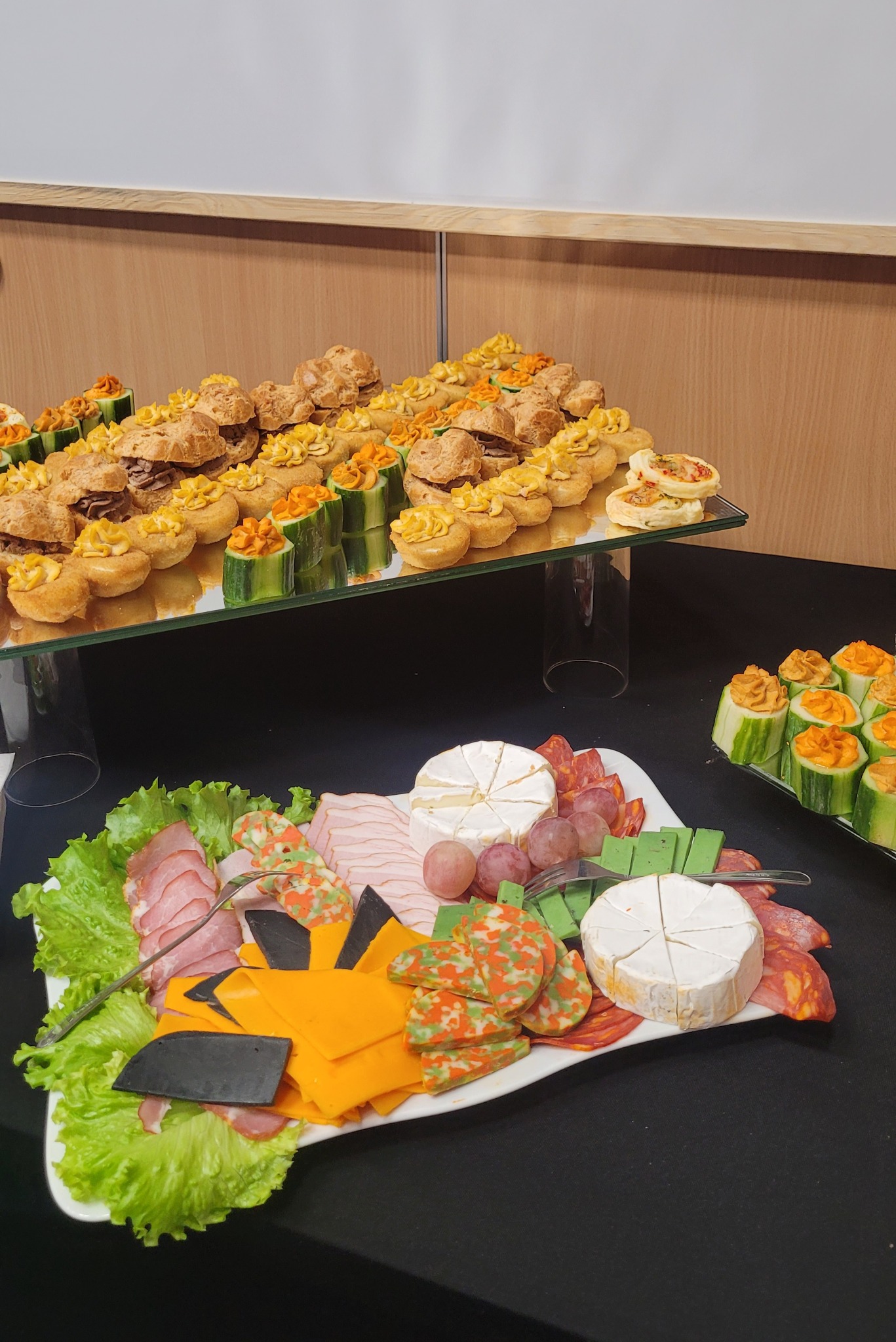 Elegancki stół cateringowy z różnorodnymi przekąskami: mini kanapeczki, koreczki z ogórka i pasty, deska serów i wędlin na czarnym obrusie. Prezentacja na dwóch poziomach.