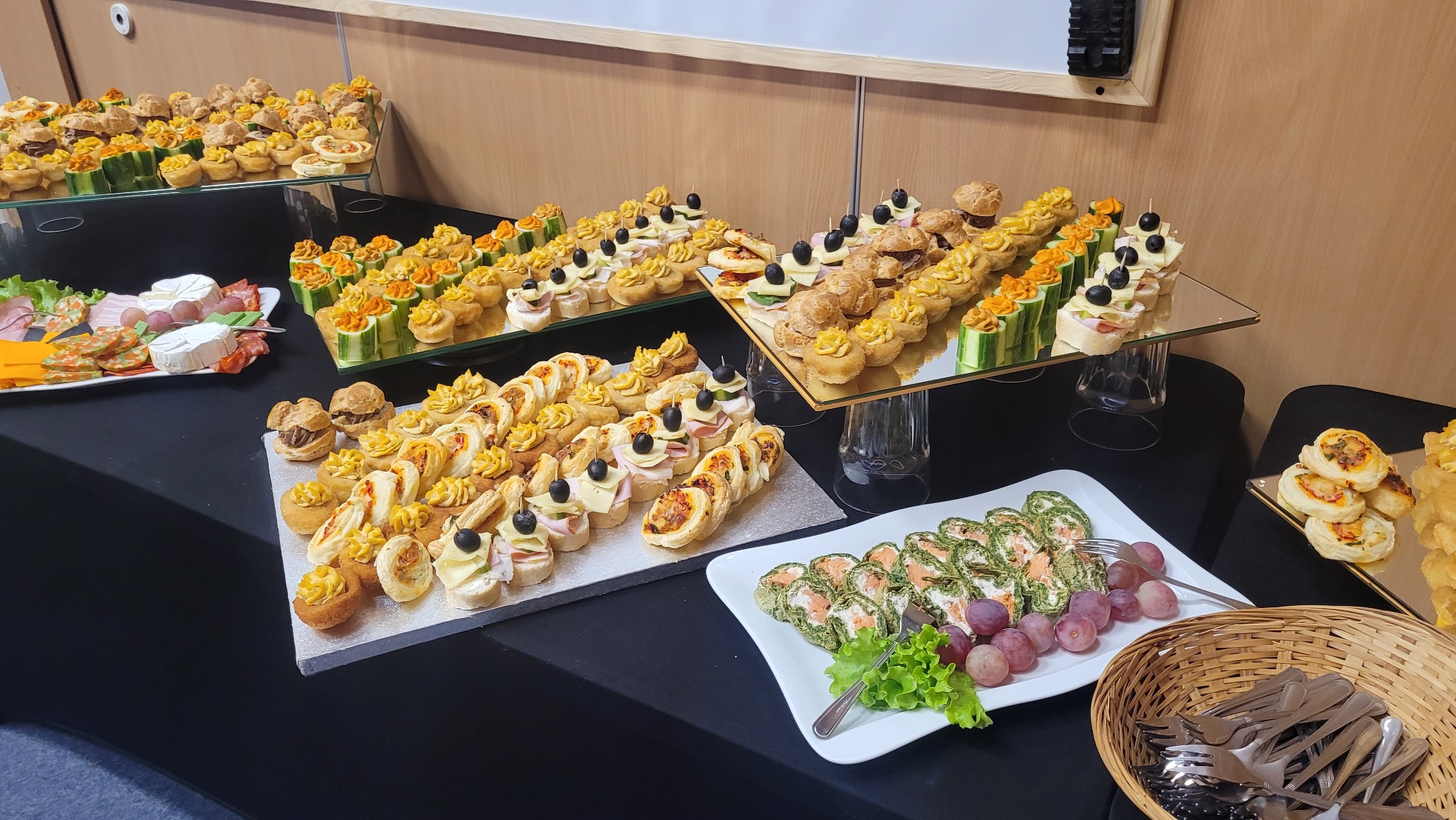 Stół cateringowy z różnorodnymi przekąskami: kanapeczki, roladki szpinakowe, koreczki, mini tartaletki i ciastka, ułożone na paterach i talerzach. Elegancki bufet.