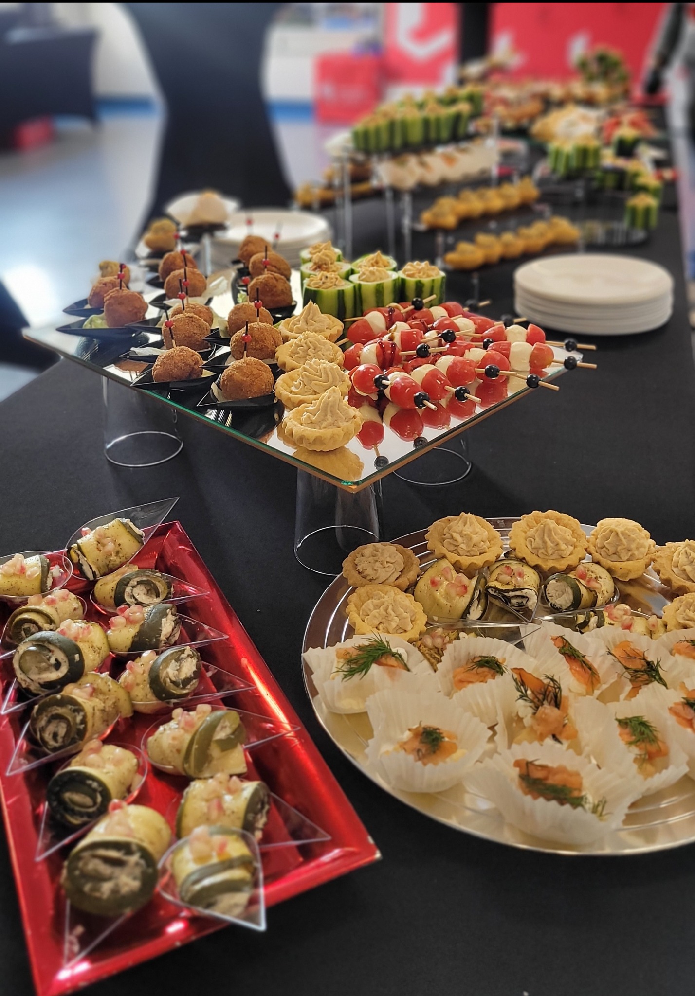 Stół cateringowy z różnorodnymi przekąskami: koreczki caprese, roladki z cukinii, tartaletki z kremem i mini babeczki z łososiem. Elegancka prezentacja na przyjęciu.
