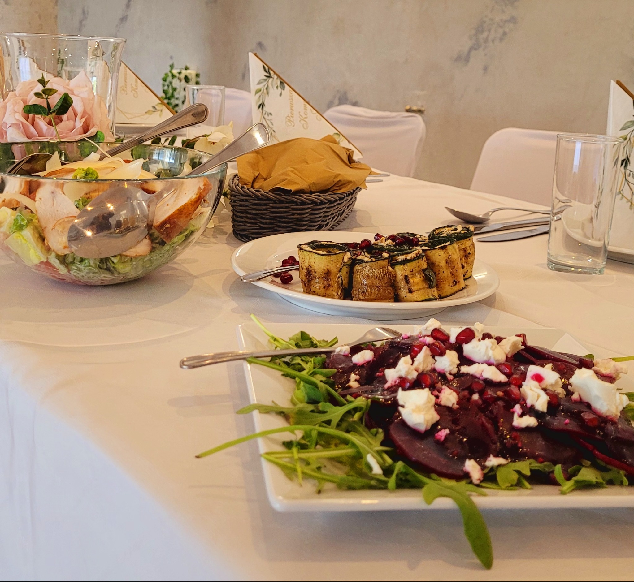 Elegancko zastawiony stół z sałatkami, roladkami z cukinii i burakami z fetą i rukolą. Dekoracje kwiatowe i serwetki z motywem roślinnym.