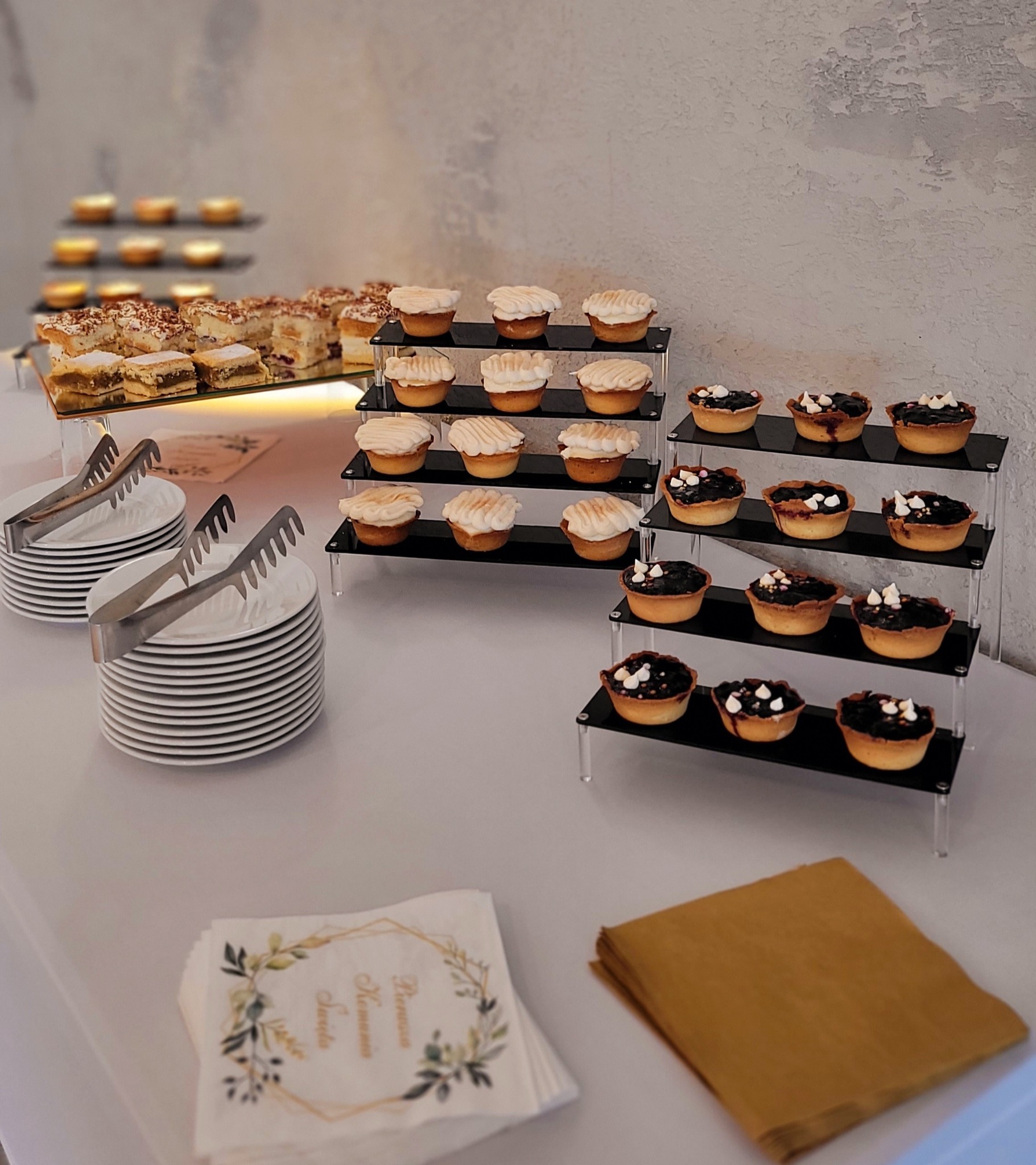 Elegancki stół z deserami: tartaletki z kremem i owocami na czarnych stojakach, ciasta, stosy talerzy i serwetki z motywem roślinnym. Catering na przyjęciu.