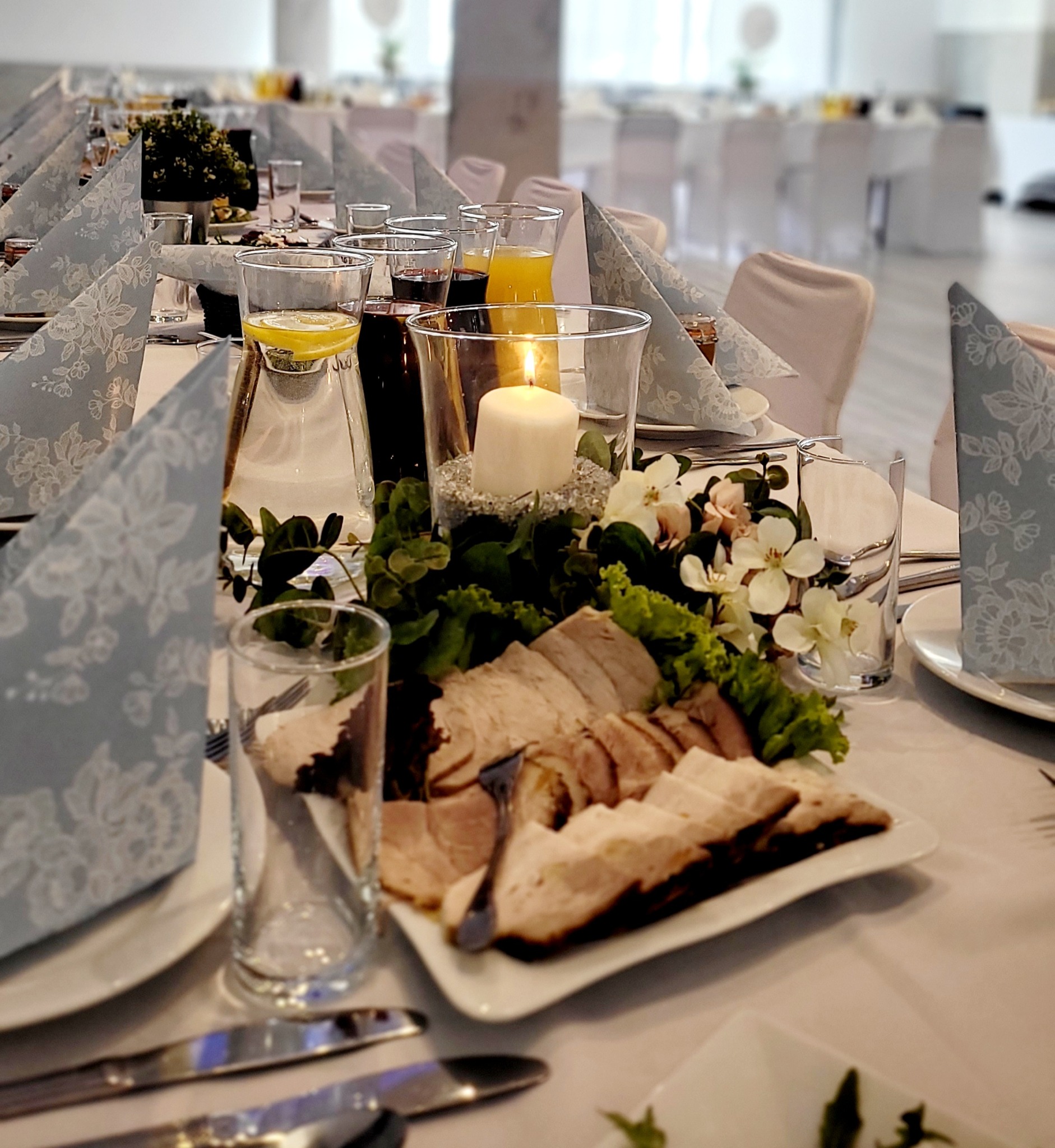Elegancko zastawiony stół z półmiskiem wędlin, świecą w szklanym naczyniu i napojami. Dekoracyjne serwetki i kwiaty dopełniają aranżację przyjęcia.