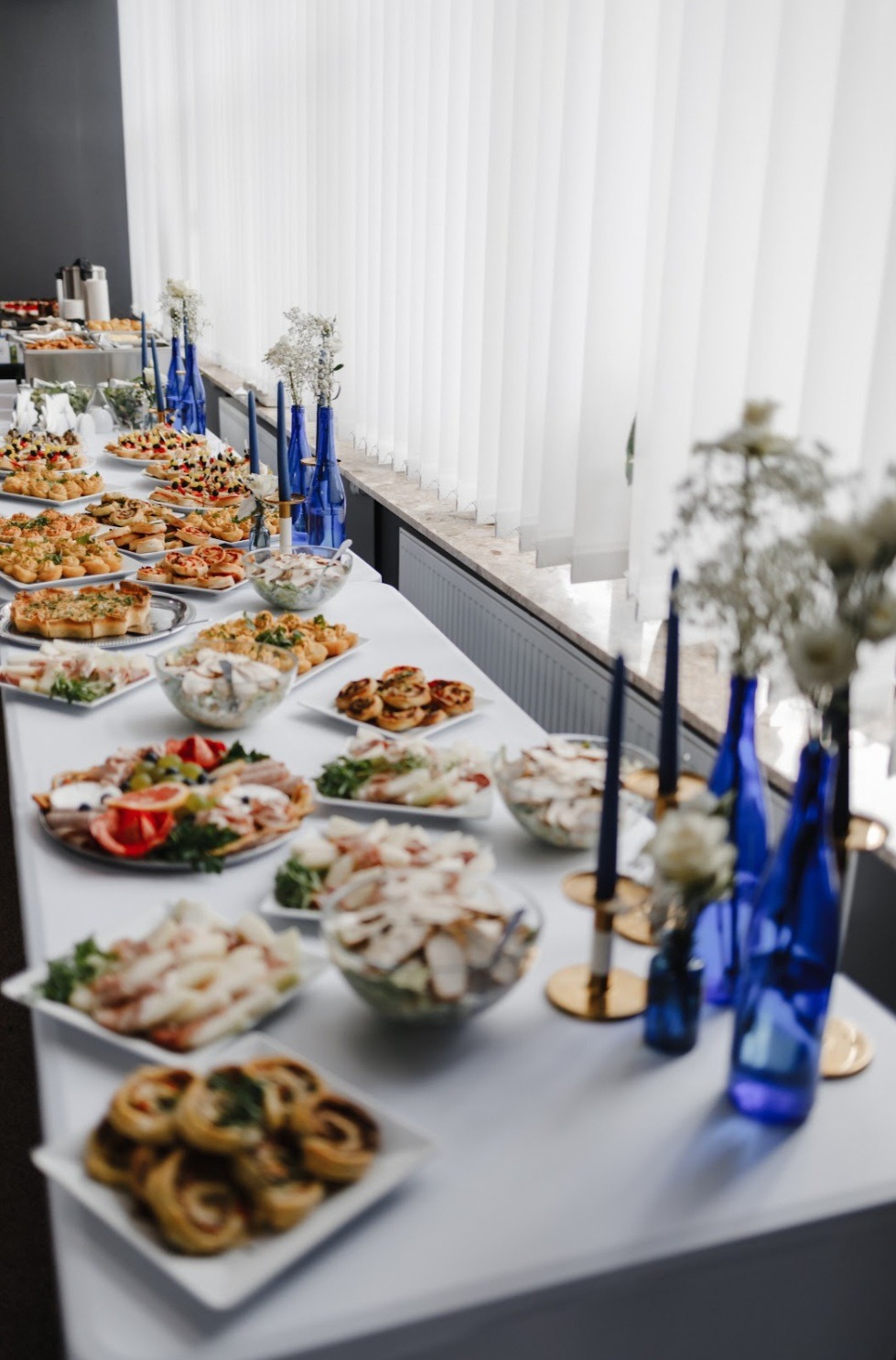 Stół cateringowy z przekąskami, sałatkami i dekoracjami w niebieskich wazonach. Różnorodność potraw, jasne tło, elegancki wystrój, widok z bliska, profesjonalna prezentacja.