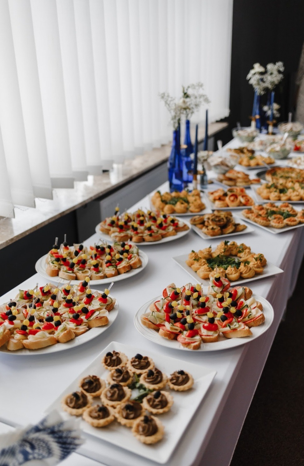 Stół cateringowy z przekąskami: kanapeczki z warzywami na wykałaczkach, tartaletki z kremem czekoladowym, białe zasłony w tle. Dekoracje z niebieskich wazonów.
