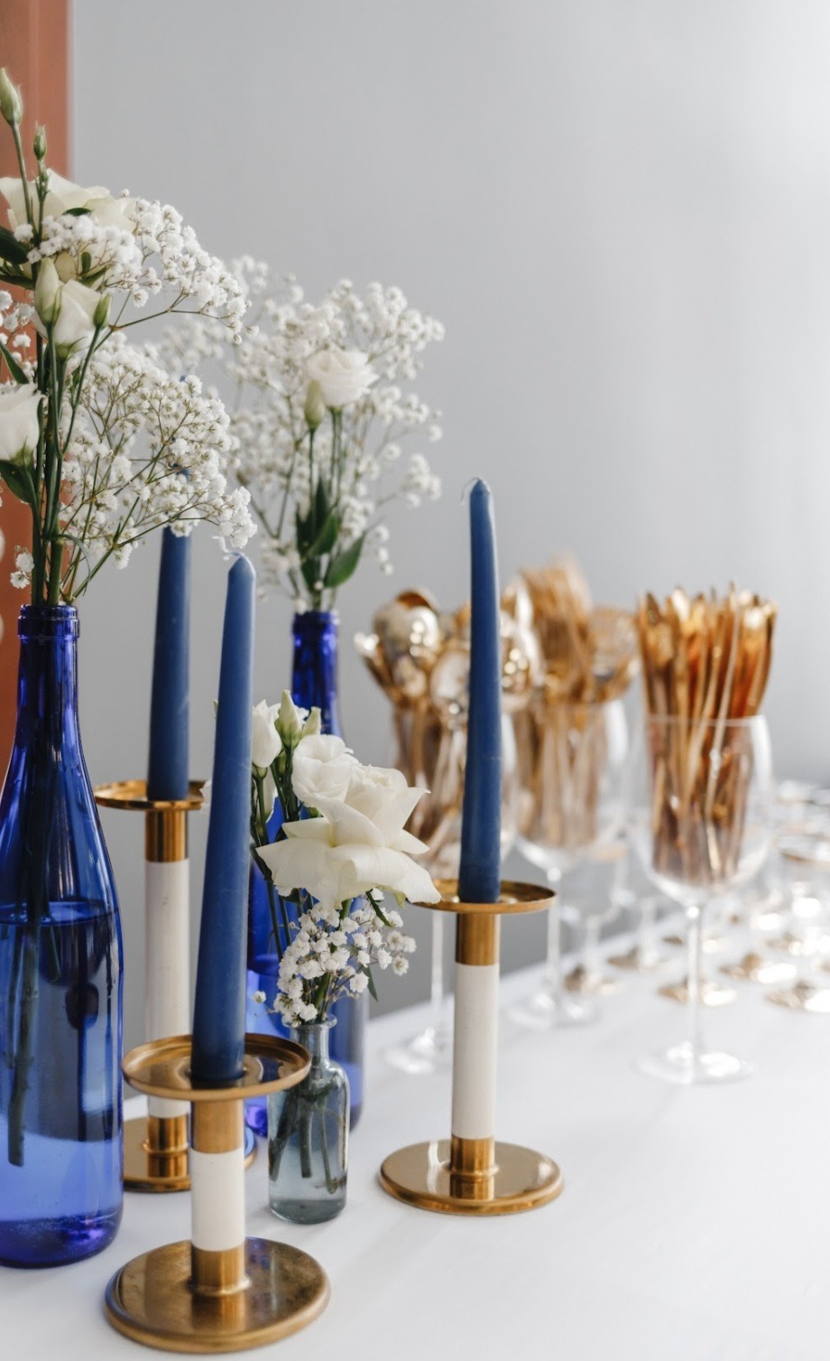 Dekoracja stołu: niebieskie świece w złotych świecznikach, kwiaty w wazonach z niebieskiego szkła, złote sztućce w kieliszkach. Elegancka aranżacja.