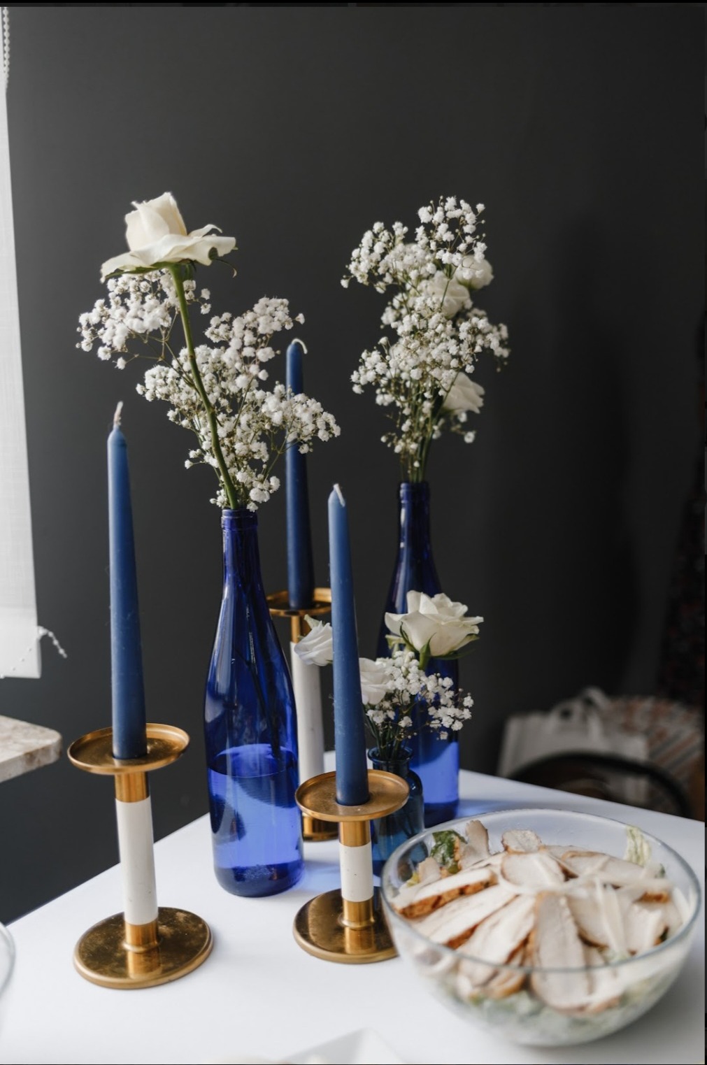 Dekoracja stołu z białymi kwiatami w niebieskich butelkach i granatowymi świecami w złotych świecznikach, obok miska z sałatką z kurczakiem na białym stole.