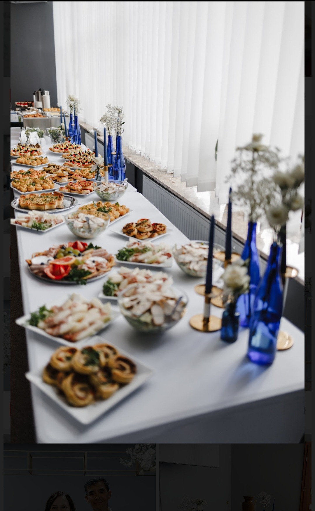 Stół cateringowy z przekąskami, sałatkami i dekoracjami w niebieskich wazonach. Długi stół przy oknie z białymi żaluzjami.