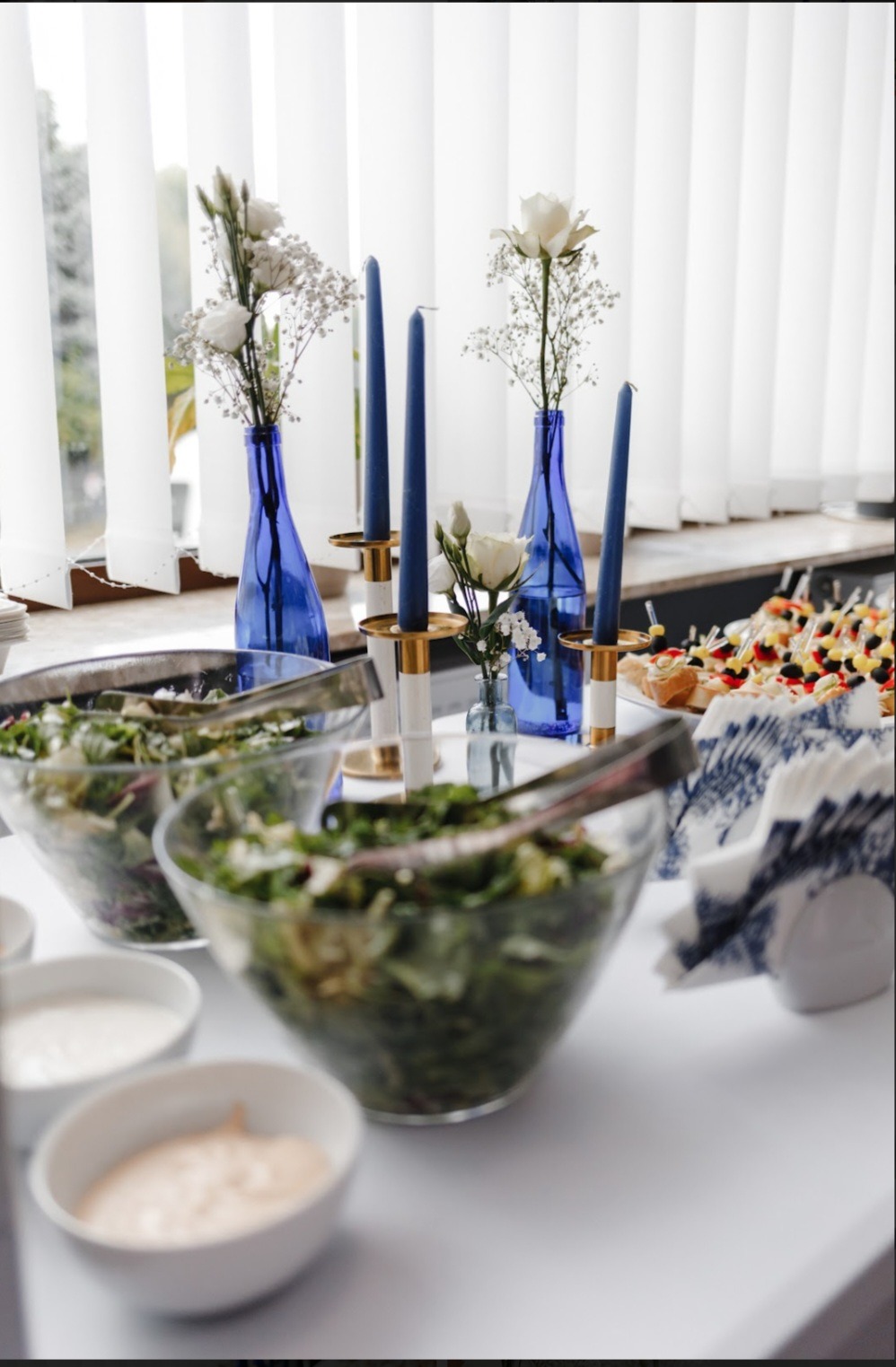 Dekoracja stołu z sałatkami, kanapeczkami, niebieskimi świecami i kwiatami w szklanych wazonach, ustawiona przy oknie z białymi żaluzjami.