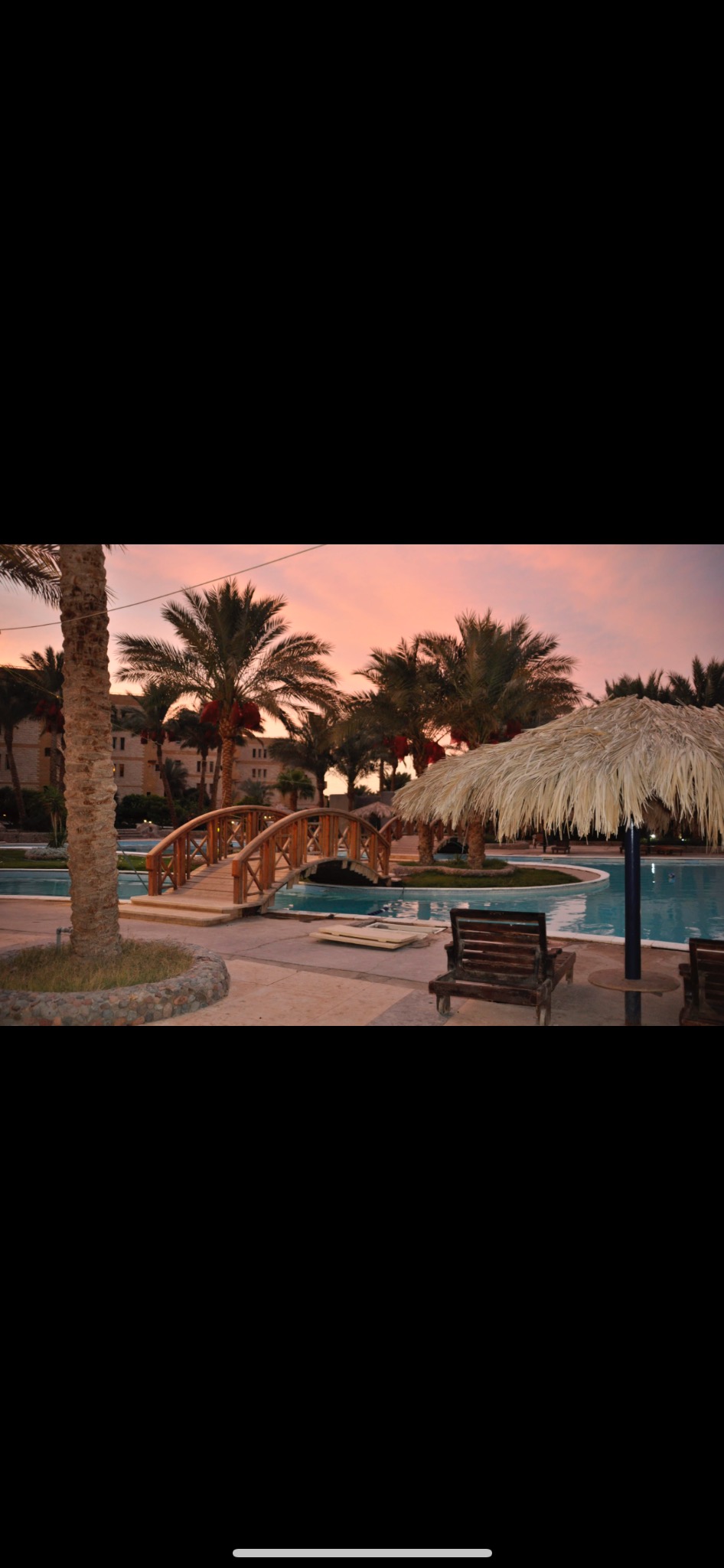 Egzotyczny basen z drewnianym mostkiem i palmami w tle o zachodzie słońca. Parasol z liści palmowych i leżaki relaksacyjne przy wodzie.