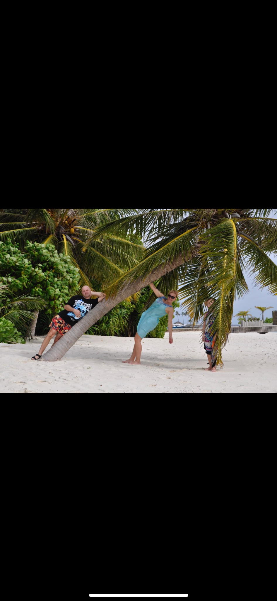 Rodzinna sesja na plaży: mężczyzna opiera się o palmę, kobieta zwisa z gałęzi, a dziecko stoi w cieniu drzewa. Biały piasek, egzotyczna roślinność w tle.