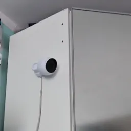 Kamera IP, biała, zamontowana na szafce w mieszkaniu w Krakowie. Widoczny kabel zasilający. Montaż kamery na wysokości, zapewniający szeroki kąt widzenia.