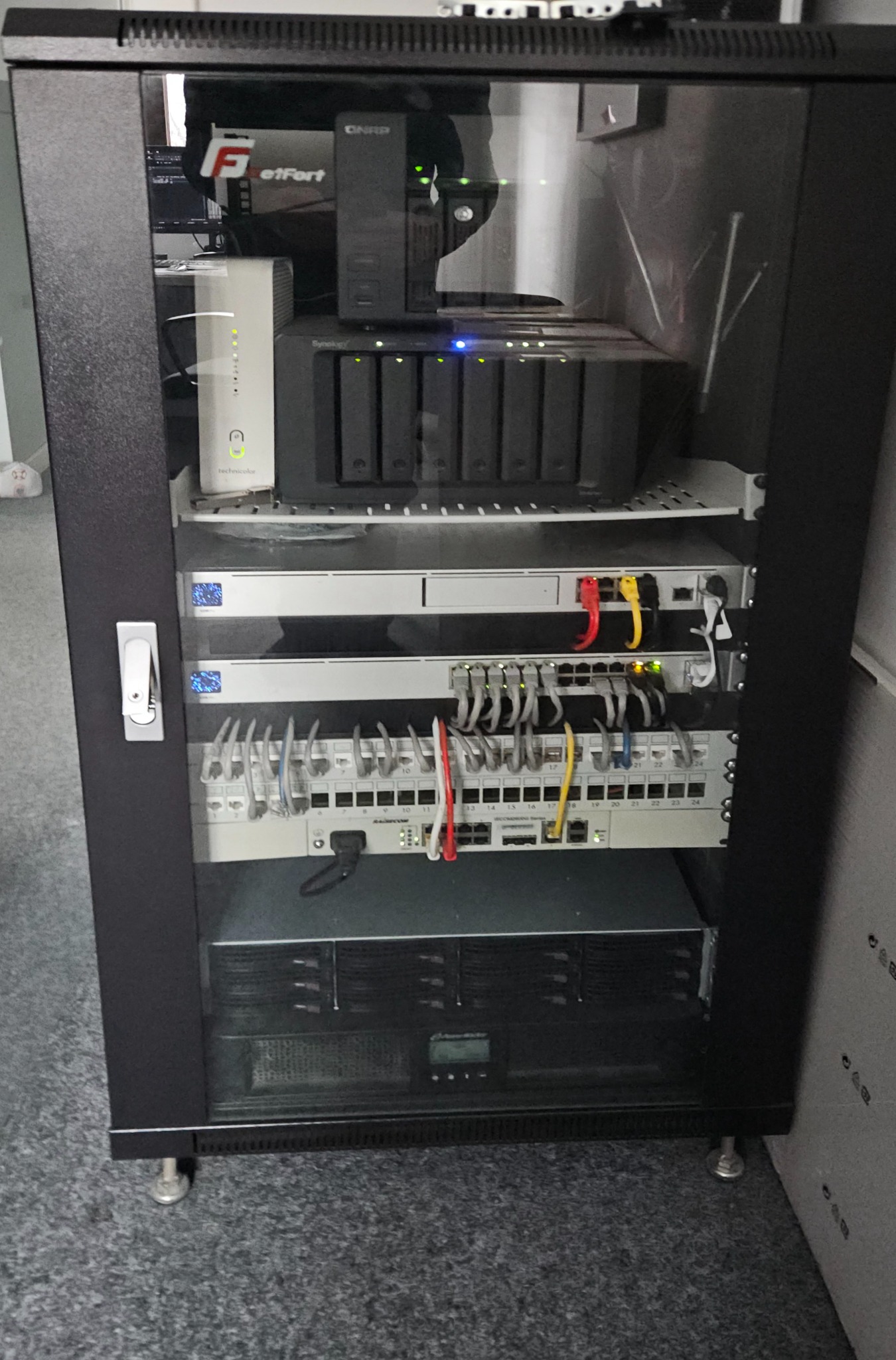 Czarna szafa rack z zainstalowanymi urządzeniami sieciowymi: serwer NAS Synology, router Technicolor, przełączniki sieciowe z podłączonymi kablami Ethernet i zasilacz awaryjny.