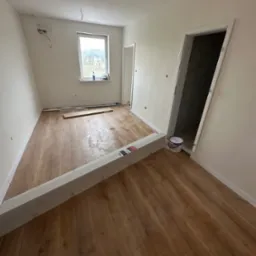 Pokój z ułożoną drewnianą podłogą, przygotowany do dalszych prac wykończeniowych. Widoczne paczki paneli, puszka z farbą i fragment okna.
