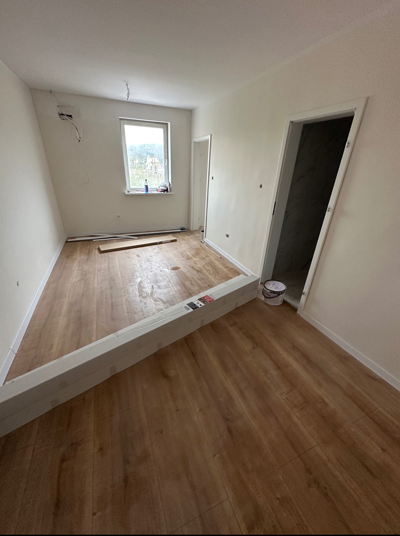 Pokój z ułożoną drewnianą podłogą, przygotowany do dalszych prac wykończeniowych. Widoczne paczki paneli, puszka z farbą i fragment okna.