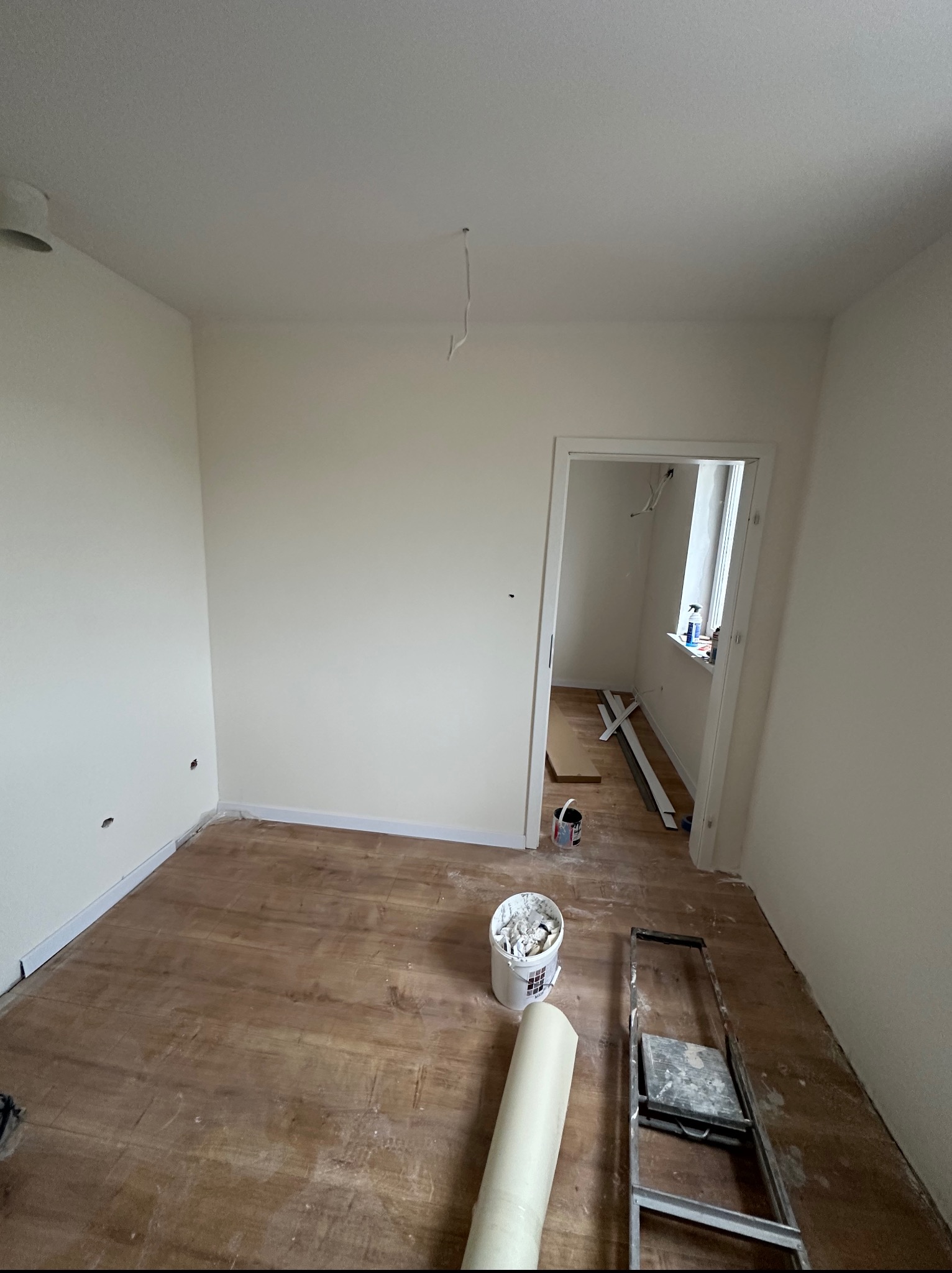 Wnętrze pokoju w trakcie odświeżania: przygotowane ściany, podłoga częściowo odsłonięta, widoczne narzędzia malarskie i drabina. Przewody oświetleniowe zwisają z sufitu.