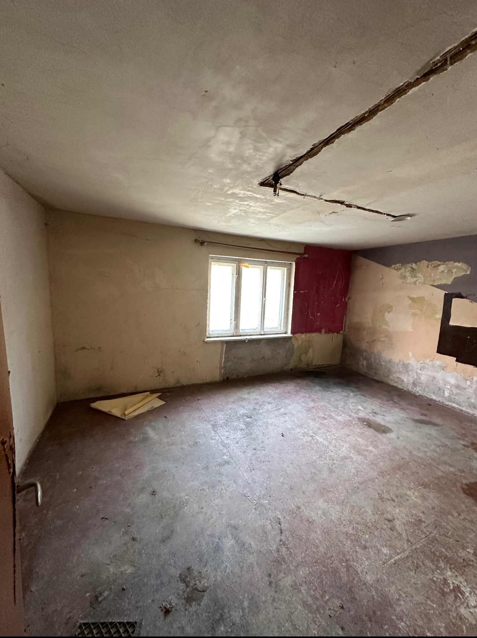 Pokój w trakcie remontu: ściany z resztkami farb, odsłonięty fragment sufitu, widoczne ślady wilgoci i zniszczeń, okno z naturalnym światłem.