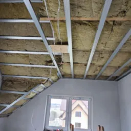 Poddasze z oknami dachowymi w trakcie ocieplania: wełna mineralna między metalowymi profilami, przygotowane do montażu płyt gipsowo-kartonowych. Widok na zewnątrz przez okno.