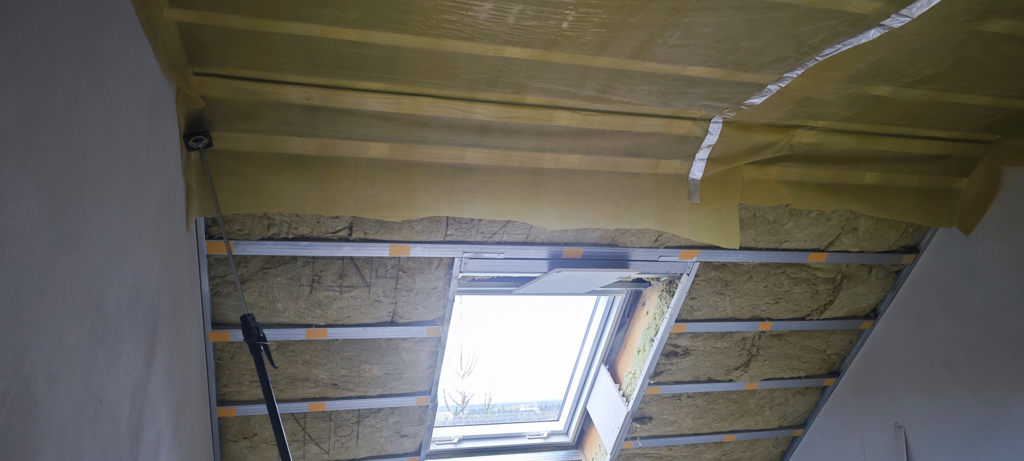 Poddasze z oknem dachowym w trakcie ocieplania wełną mineralną, widoczne profile metalowe i folia paroizolacyjna. Prace wykończeniowe.