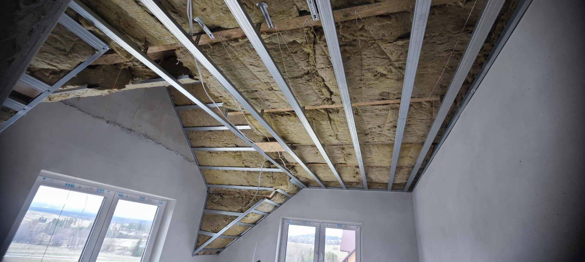 Poddasze z izolacją z wełny mineralnej i metalowym stelażem pod płyty gipsowe, widok z dołu, okna w tle. Ocieplenie skośnego stropu.