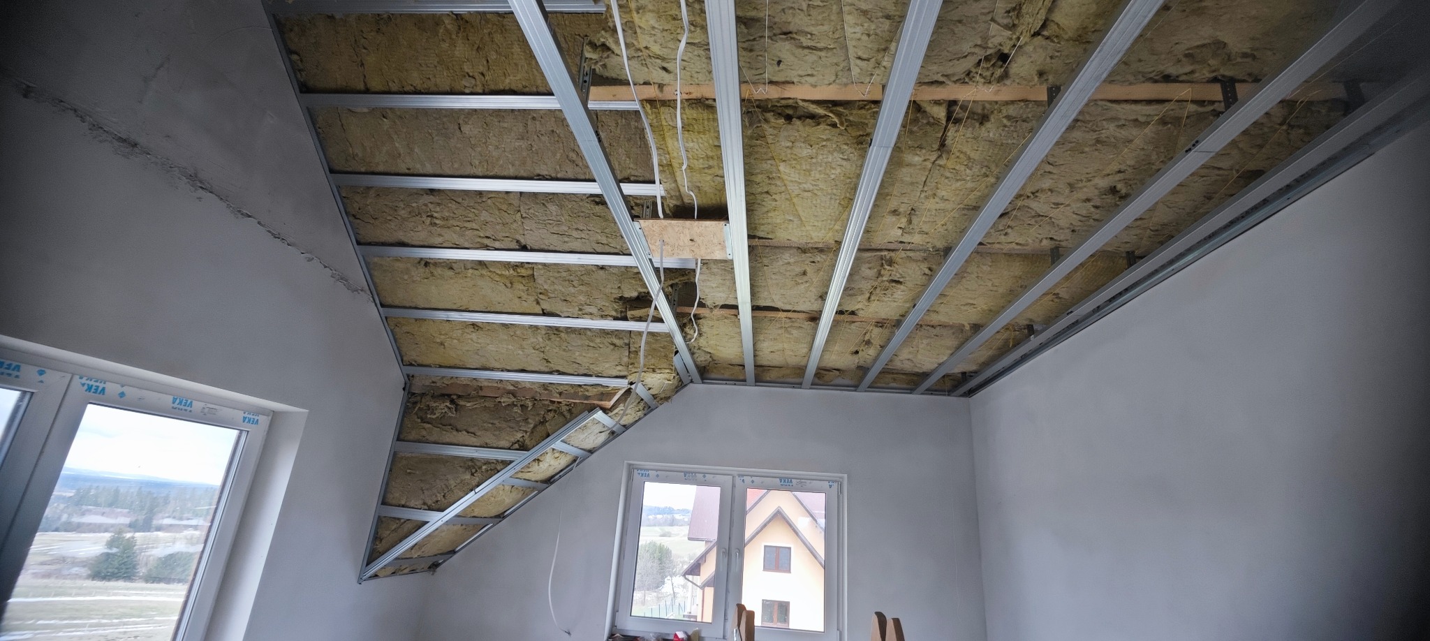 Poddasze z oknami dachowymi w trakcie ocieplania: wełna mineralna między metalowymi profilami, przygotowane do montażu płyt gipsowo-kartonowych. Widok na zewnątrz przez okno.