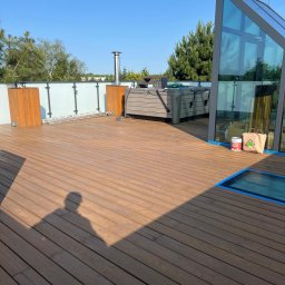 RHINP SPÓŁKA Z OGRANICZONĄ ODPOWIEDZIALNOŚCIĄ - Taras z drewna po czyszczeniu w Poznaniu. Widoczne akcesoria do impregnacji i cień osoby wykonującej usługę. W tle jacuzzi i elementy architektury.