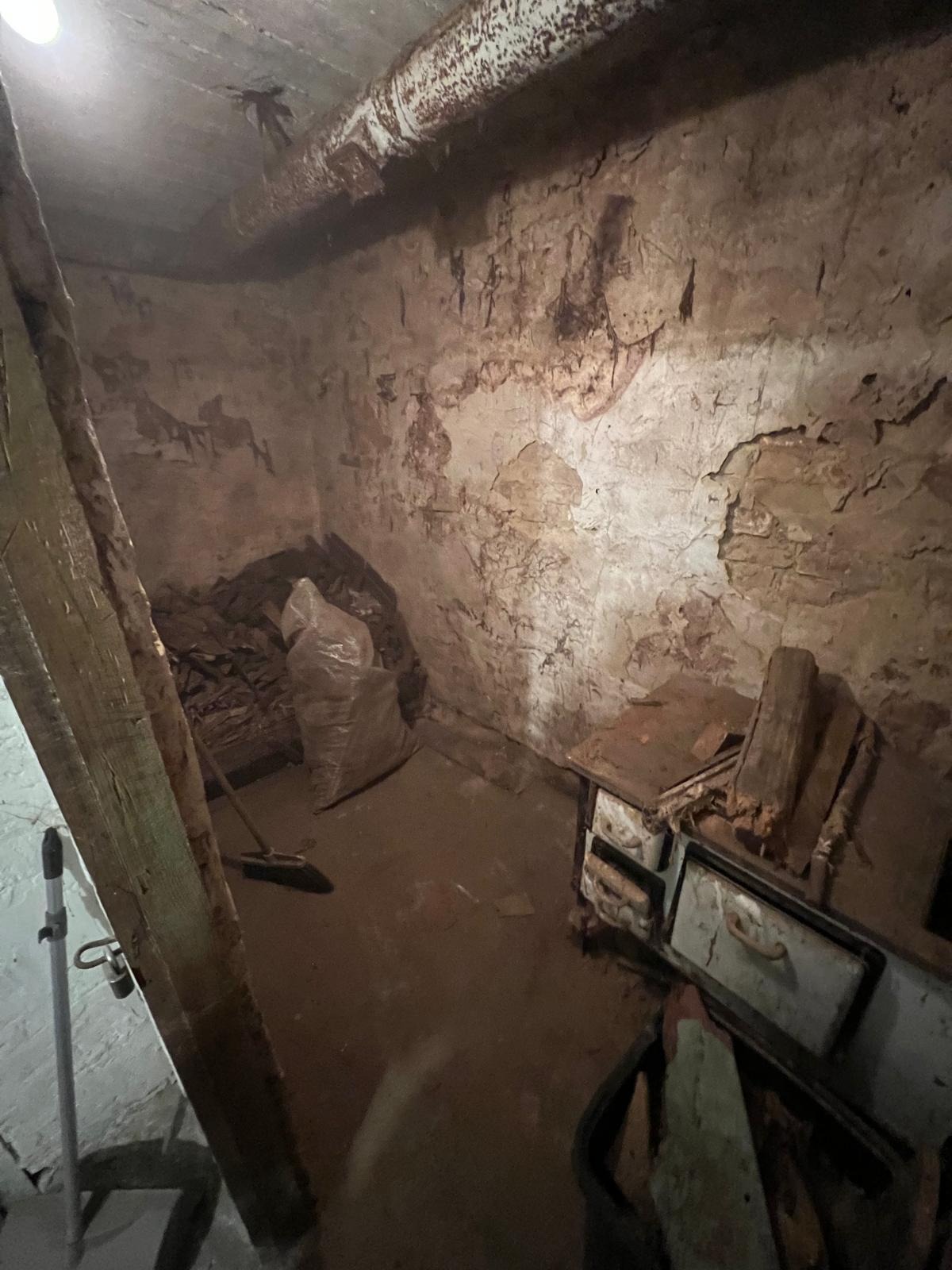 Zaniedbane pomieszczenie piwniczne z odpadami, starą kuchenką i śladami wilgoci na ścianach. Widoczna rura pod sufitem i słabe oświetlenie.