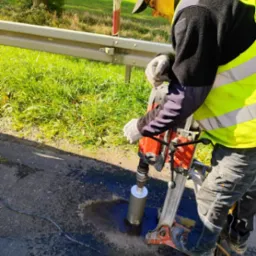 Pracownik w kamizelce wierci rdzeniówką otwór w asfalcie na drodze, obok barierka i trawa. Widoczne narzędzia i etap pracy.