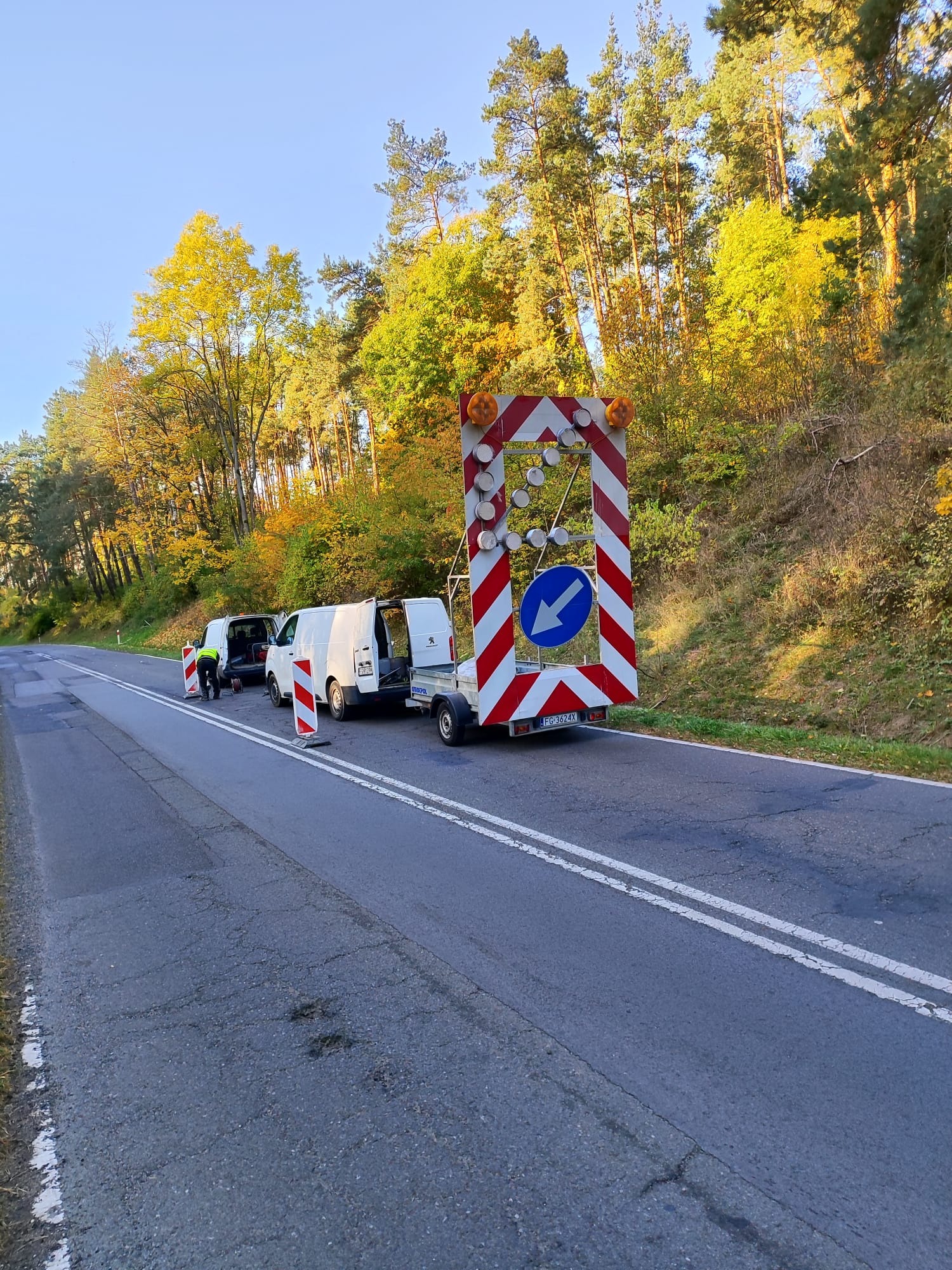Drogowcy przy ustawianiu znaku objazdu na uszkodzonej drodze, w tle jesienny las i dwa białe busy. Praca wykonywana w słoneczny dzień.
