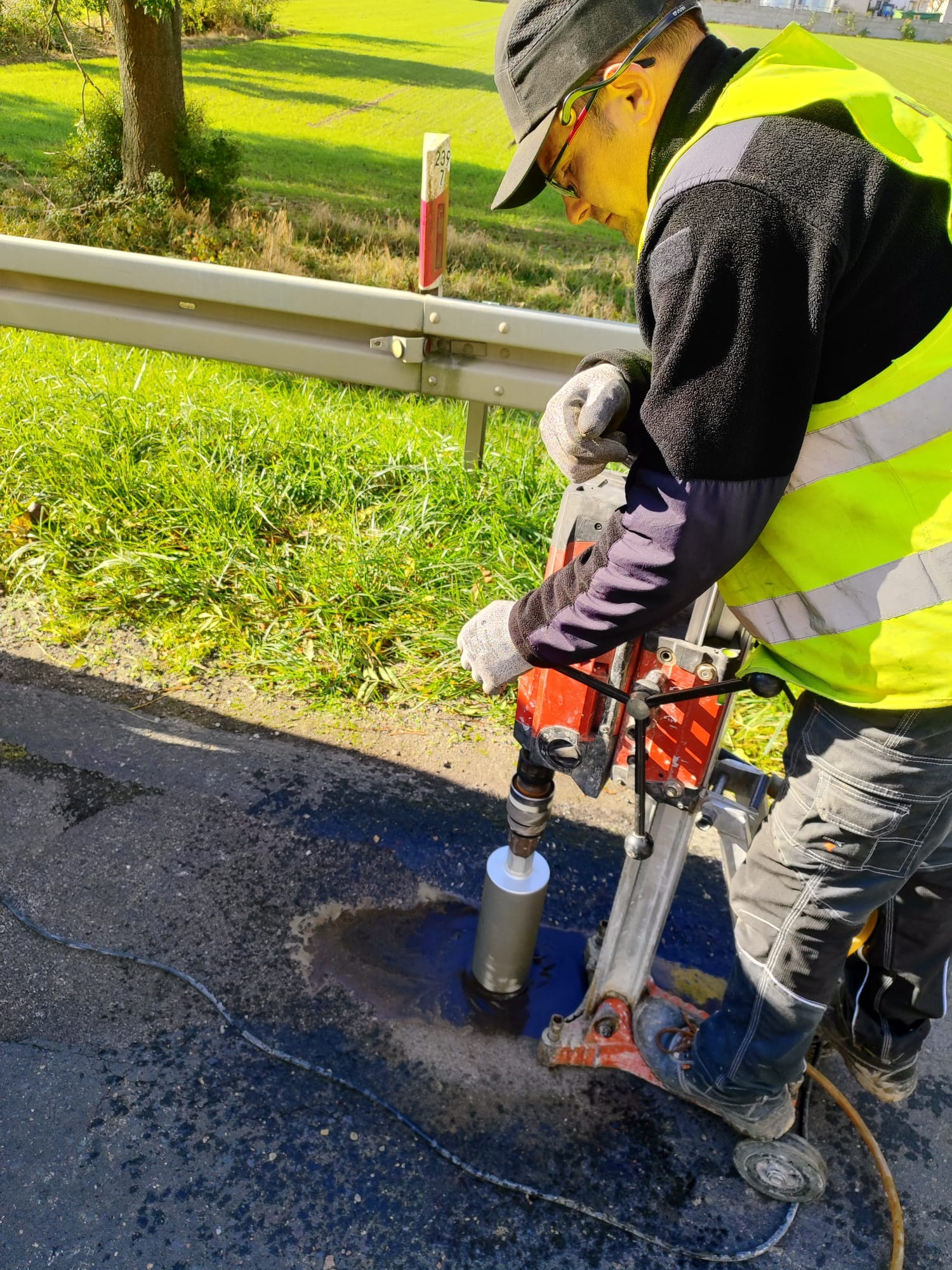 Pracownik w kamizelce wierci rdzeniówką otwór w asfalcie na drodze, obok barierka i trawa. Widoczne narzędzia i etap pracy.