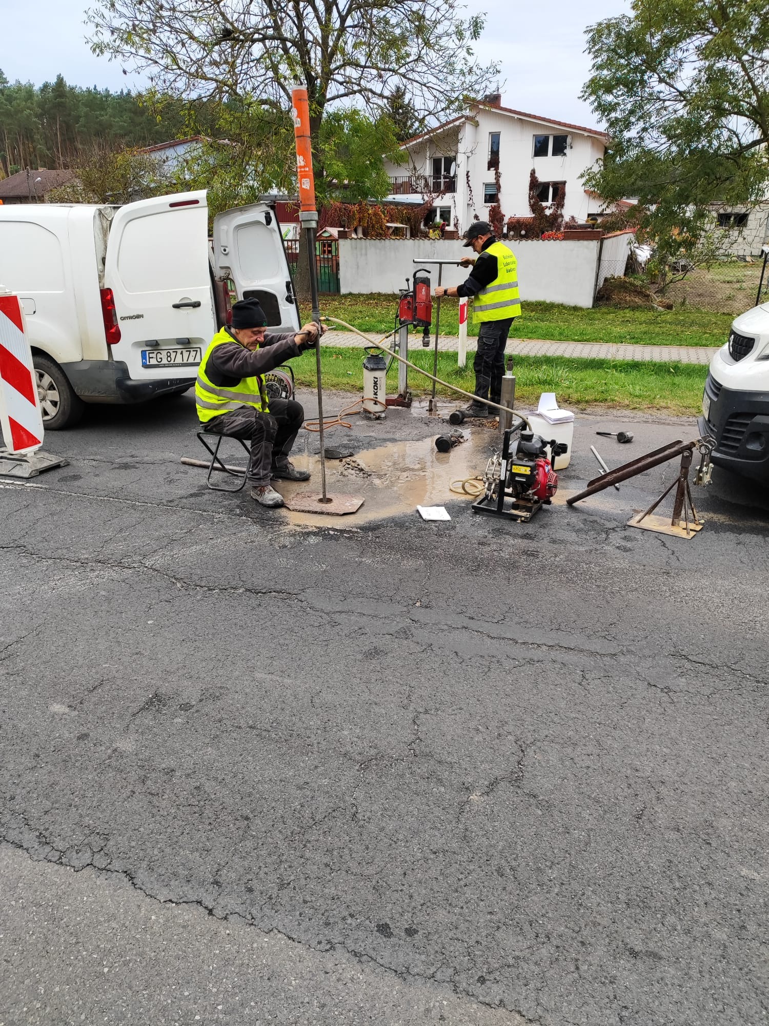 Prace geologiczne na drodze: dwóch mężczyzn w kamizelkach wierci otwór w asfalcie. Sprzęt w pobliżu, białe samochody w tle. Wiercenie w asfalcie.